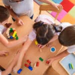 foto dall'altro di 5 bambini che giocano sun un banco con dei colori, sullo sfondo un tappeto a quadrati collorati
